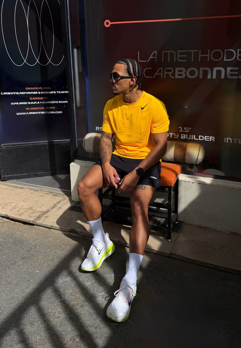 Homme aux cheveux tressés, portant un t-shirt jaune, un short noir et des baskets blanches, assis sur un banc devant un studio de fitness sous la lumière du soleil.