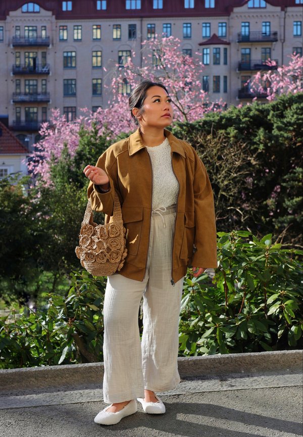 Brun mockajacka i oversized-stil, vit texturerad topp, beige linnbyxor, vita fluffiga skor och en vävd väska med blommönster, mot en bakgrund av träd.