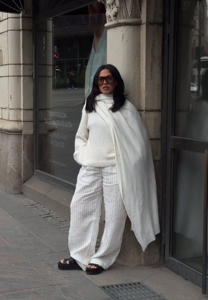 Jersey blanco, bufanda blanca drapeada y pantalones de pierna ancha a rayas. Sandalias de plataforma negras aportan contraste. Entorno urbano con paredes de piedra.