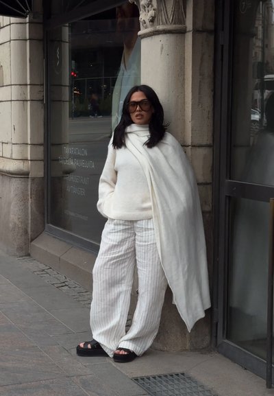 Jersey blanco, bufanda blanca drapeada y pantalones de pierna ancha a rayas. Sandalias de plataforma negras aportan contraste. Entorno urbano con paredes de piedra.