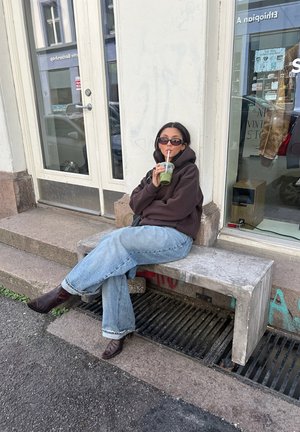 Una persona está sentada en un banco de concreto gris, vistiendo una sudadera marrón y jeans de mezclilla azul claro, sosteniendo una bebida verde con una pajita.