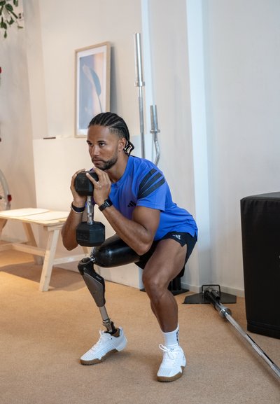 Una mancuerna negra sostenida con ambas manos por una persona con una camiseta deportiva azul y pantalones cortos negros, en cuclillas sobre un suelo beige, con una pierna protésica.