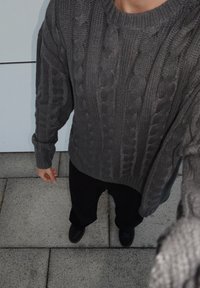 Überdimensionierter grauer Kabelstrickpullover mit gerippten Bündchen; kombiniert mit schwarzen, weiten Hosen und schwarzen Schuhen. Strukturiertes Strickmuster sichtbar.