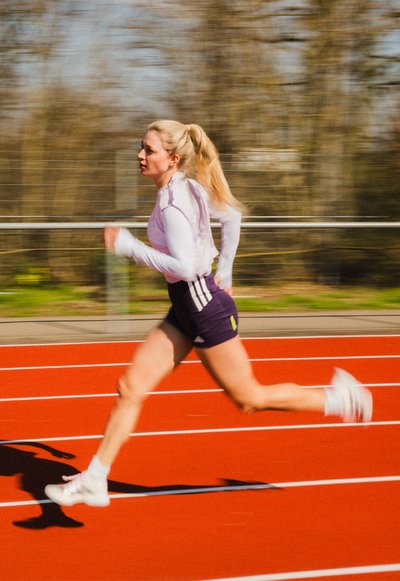 Mujer con cabello rubio recogido en una coleta corriendo con pantalones cortos morados y camiseta blanca de manga larga en una pista al aire libre roja durante el día.