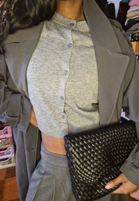 Sieviete valkā pelēku īso pogājamo kardiganu, atbilstošas augsta vidukļa bikses, tumši pelēku mēteļa virskārtu un tur melnu austu rokassomu.