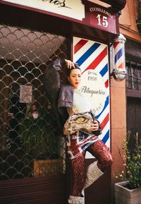 Model in einer dunklen, übergroßen Jacke, grafischem Kleid mit Union Jack, gemusterten roten Strumpfhosen und metallischen kniehohen Stiefeln, hält eine Tasche mit Reptilienmotiv.