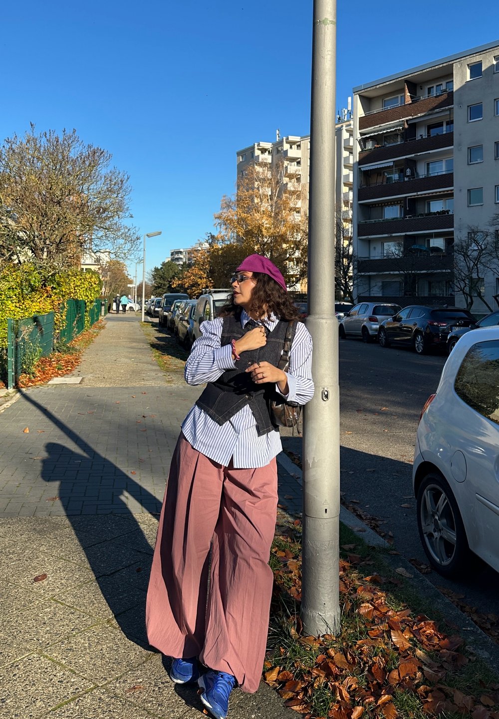 Model purtând o cămașă în dungi albastre și albe, vestă neagră și pantaloni largi roz, sprijinită de un stâlp pe stradă într-un decor urban.