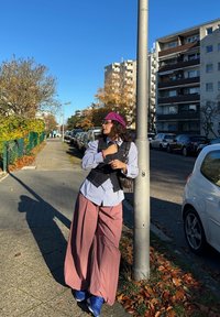 Model purtând o cămașă în dungi albastre și albe, vestă neagră și pantaloni largi roz, sprijinită de un stâlp pe stradă într-un decor urban.