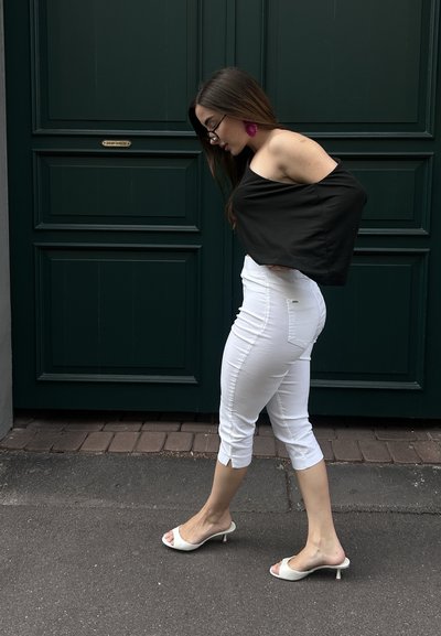 Top negro sin mangas, pantalones blancos recortados con cortes laterales. Mules blancos de tacón. De pie contra una puerta verde.