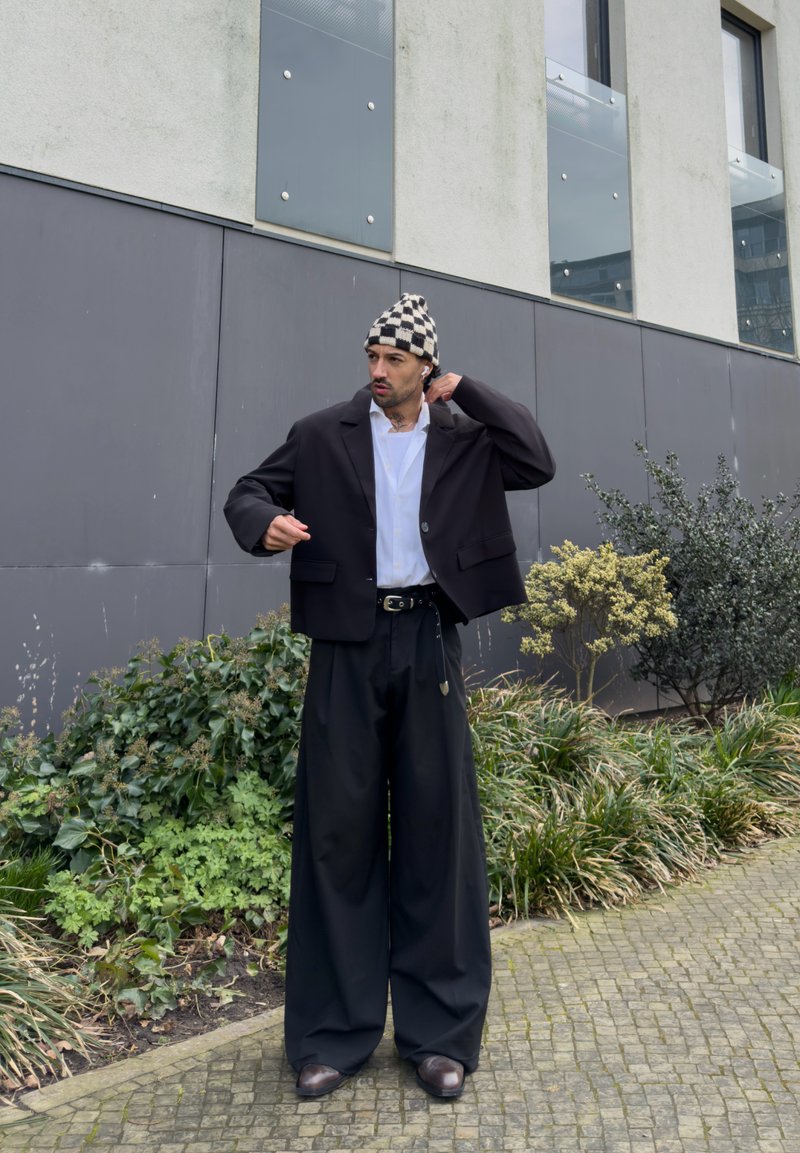 Homme portant une veste de costume noire, un pantalon noir à jambes larges, une chemise blanche, un bonnet à carreaux et des écouteurs, debout à l'extérieur près d'un bâtiment et de la verdure.