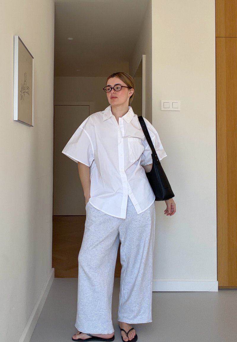 Femme portant une chemise blanche boutonnée, un pantalon large gris, des lunettes noires et un sac à épaule noir, debout dans un couloir minimaliste.