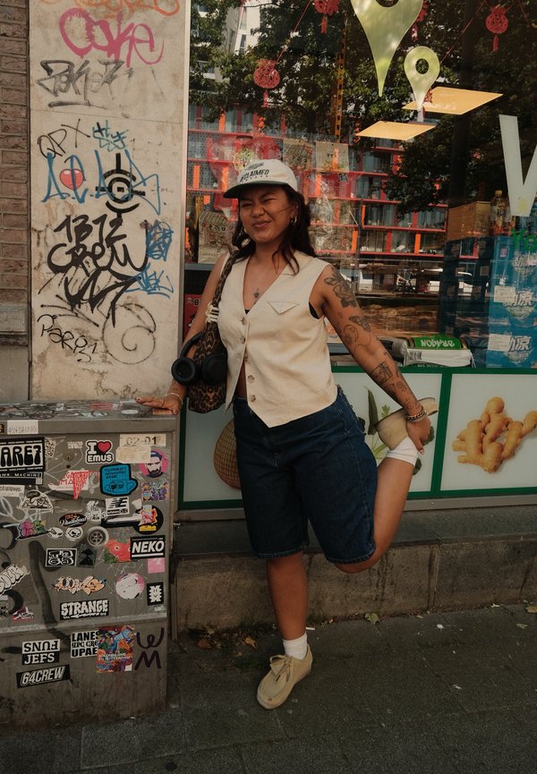 Gilet senza maniche beige, pantaloncini in denim blu e scarpe color crema. Presenta tatuaggi e una borsa con stampa leopardata. Lo sfondo include graffiti e vetrine di negozi.