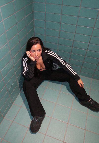 Pantalones deportivos negros y chaqueta con cremallera con rayas blancas; zapatos negros con un detalle decorativo; sobre un fondo de pared con azulejos verde azulado.