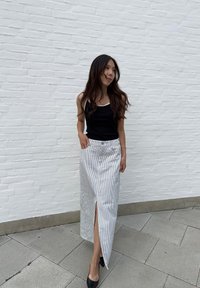 Ριγέ μακριά φούστα σε λευκό και μπλε με σκίσιμο μέχρι τον μηρό, φορεμένη με μαύρη αμάνικη μπλούζα και μαύρες γόβες μπροστά από έναν λευκό τοίχο.