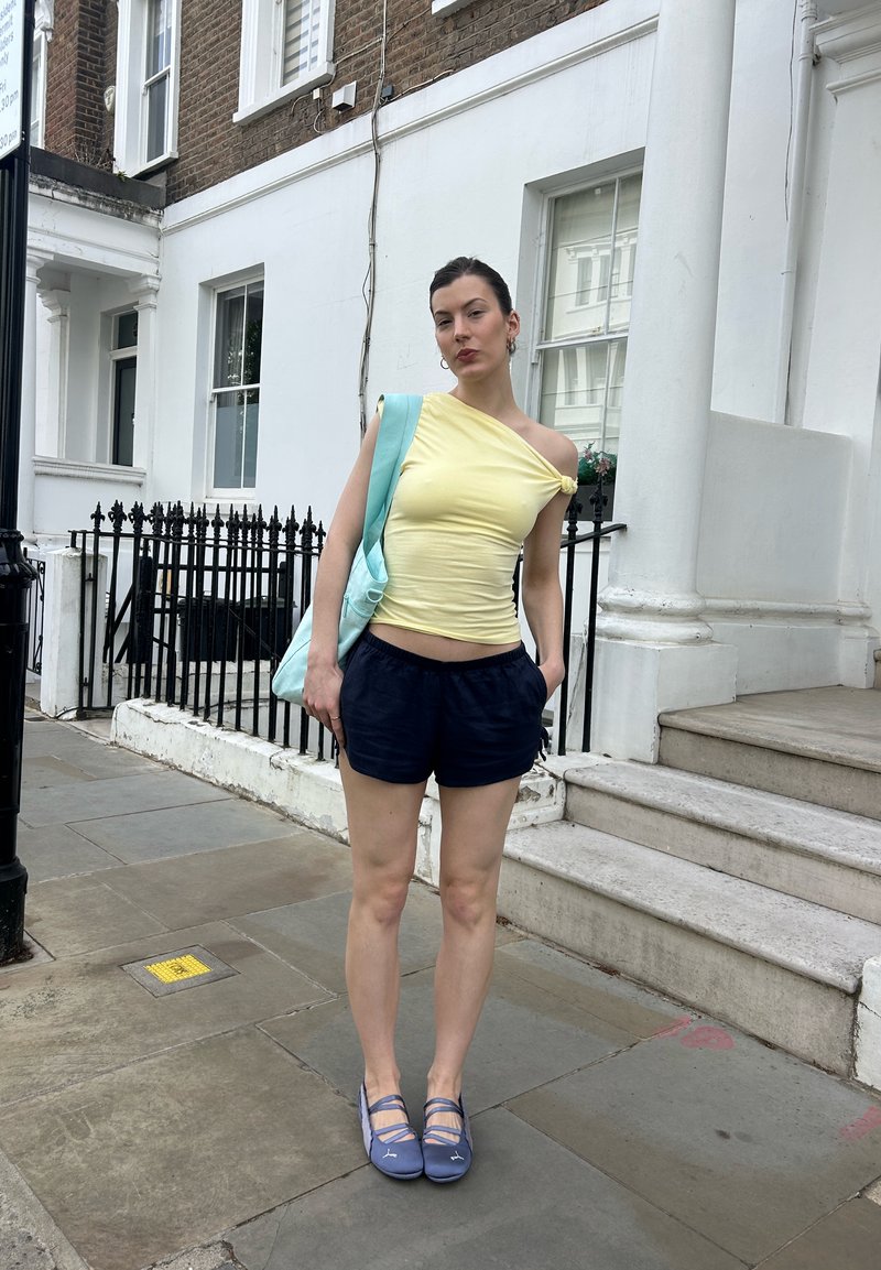 Femme debout sur un trottoir en ville, portant un haut jaune à une épaule, un short bleu marine, des chaussures bleues, et portant un sac fourre-tout bleu clair sur l'épaule.
