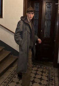 Manteau trench gris avec capuche, superposé sur un haut marron et un pantalon beige. Portes en bois foncé et sol en carreaux à motifs en arrière-plan.