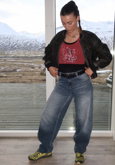 Chaqueta bomber negra, camiseta sin mangas gráfica roja, jeans amplios azules, cinturón negro. El calzado presenta detalles en amarillo y negro. Entorno interior.