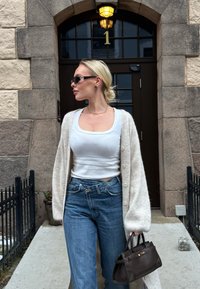 Femme blonde portant des lunettes de soleil, un débardeur blanc, un cardigan beige, un jean bleu, tenant un sac à main foncé, debout près de l'entrée en pierre d'un bâtiment numéroté 1.