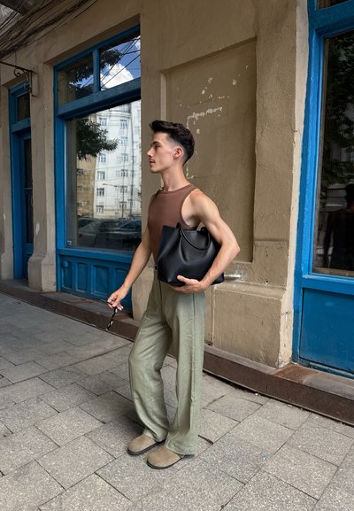 Joven con cabello corto y oscuro, vistiendo una camiseta sin mangas marrón y pantalones verdes, sosteniendo una bolsa negra y gafas de sol junto a una pared beige desgastada.