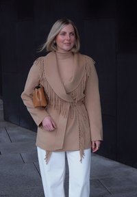 Manteau beige avec des détails à franges, design enveloppant et manches longues. Associé à un pantalon blanc et un petit sac à main marron. Texture lisse.