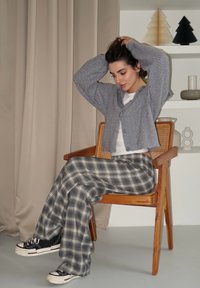 Cardigã cinza texturizado sobre uma camisa branca, combinado com calças largas de xadrez azul e cinza e ténis negros, sentado numa cadeira de madeira.