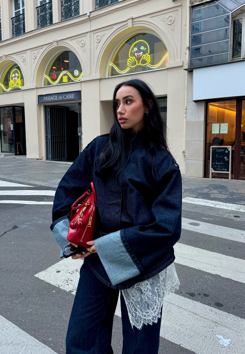 Giacca di jeans con maniche ampie, abbinata a pantaloni di jeans. Dettaglio in pizzo all'orlo e borsa rossa con rifiniture dorate. Sfondo urbano di strada.