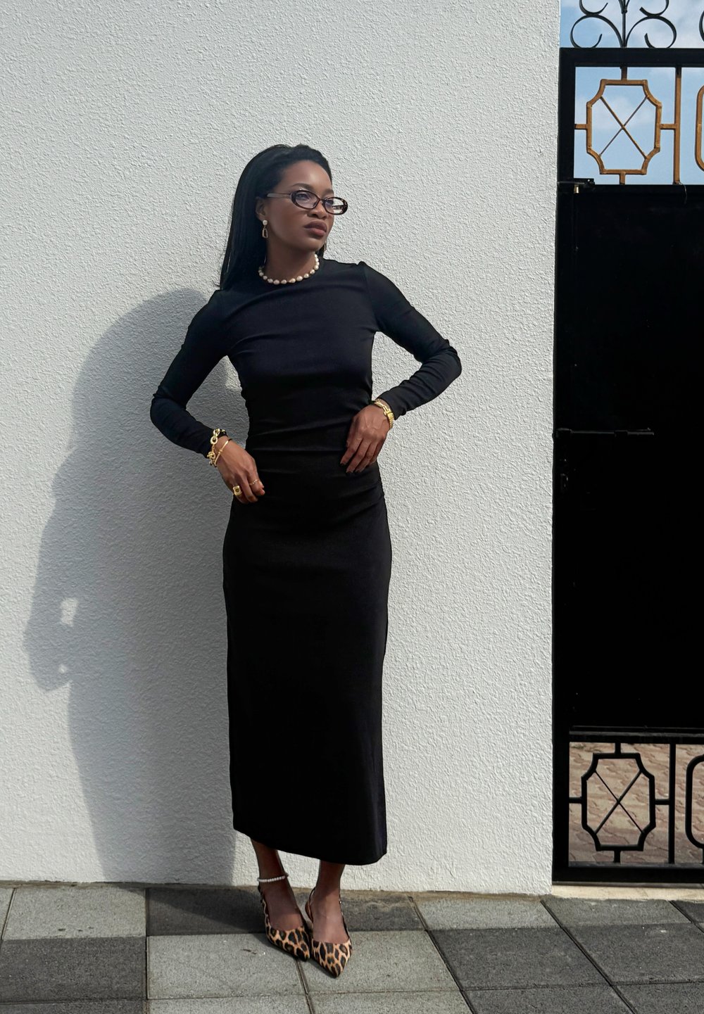 Black long-sleeve fitted dress with a straight silhouette. Accessories include a pearl necklace and gold bracelets. Leopard-print high heels.