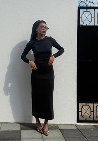 Black long-sleeve fitted dress with a straight silhouette. Accessories include a pearl necklace and gold bracelets. Leopard-print high heels.