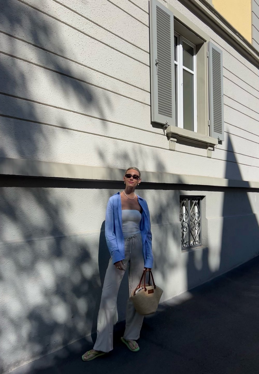 Camisa riscada azul clara sobre um top tubo branco, calças bege e uma bolsa de palha, em pé contra uma parede texturizada branca com sombras.