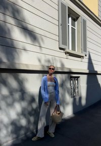 Camisa riscada azul clara sobre um top tubo branco, calças bege e uma bolsa de palha, em pé contra uma parede texturizada branca com sombras.