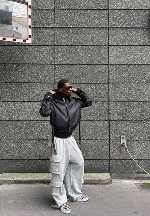 Chaqueta bomber de cuero negro con un diseño recortado, pantalones de chándal grises con bolsillos cargo y zapatos de lona de cuadros, sobre una pared gris texturizada.