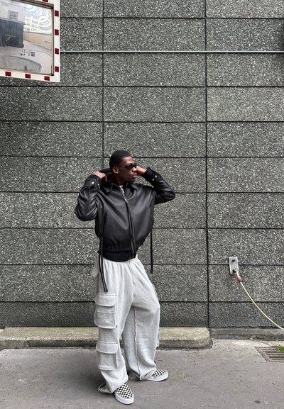 Chaqueta bomber de cuero negro con un diseño recortado, pantalones de chándal grises con bolsillos cargo y zapatos de lona de cuadros, sobre una pared gris texturizada.