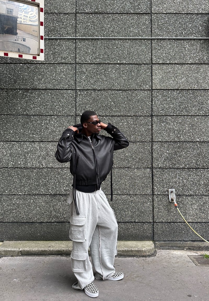 Schwarze Leder-Bomberjacke mit einem cropped Design, graue Sweatpants mit Cargo-Taschen und karierte Slipper auf einer strukturierten grauen Wand.