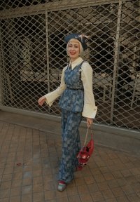 Persona sonriendo y caminando, vestida con un chaleco y pantalones de cuadros azules, blusa crema, sombrero negro con lazo, sosteniendo un bolso rojo con un charm de peluche.