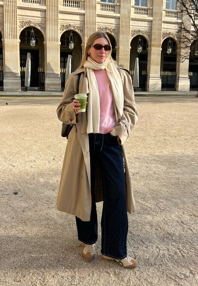 Trench beige sobre un suéter rosa, jeans de pierna ancha azul marino y zapatillas beige con acentos verdes. Sosteniendo una bebida verde. Lleva gafas de sol.