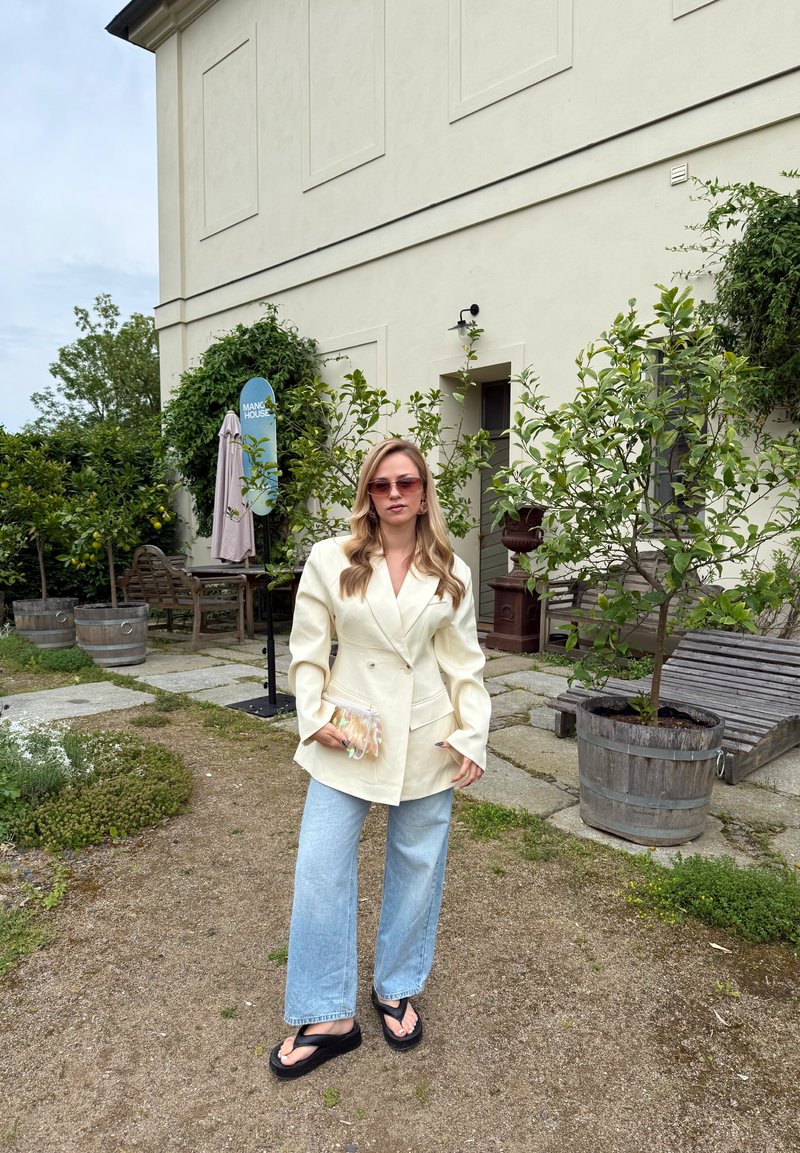 Femme portant un blazer crème, un jean bleu clair et des sandales noires, debout sur un chemin de jardin près de citronniers en pot et d’un bâtiment beige.