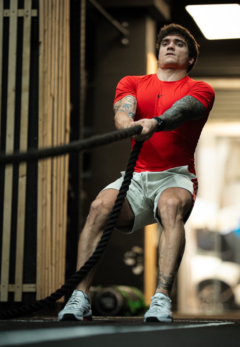 Jeune homme avec des tatouages tire une corde lourde en étant accroupi dans une salle de sport, portant un t-shirt rouge, un short blanc et des baskets blanches.