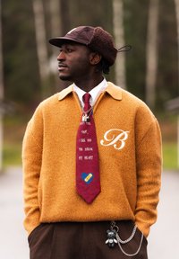 Homme portant un chapeau marron, un pull moutarde avec un "B" blanc, une cravate rouge avec du texte et un cœur, un pantalon marron avec un porte-clés ours, en extérieur dans une forêt floue.
