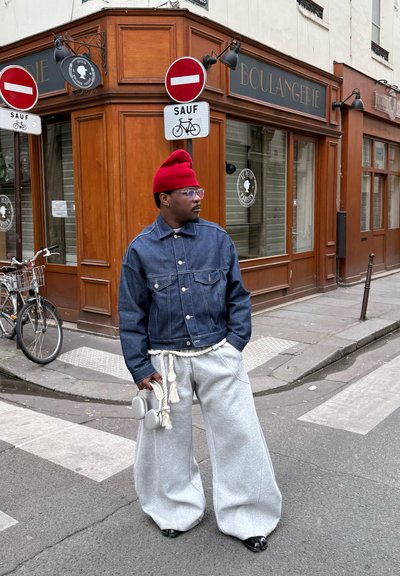 Chaqueta de mezclilla con bolsillos en el pecho, pantalones de chándal grises de pierna ancha con cordón, gorro rojo y gafas de gran tamaño, de pie en una calle.