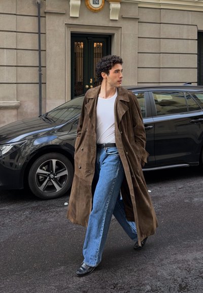 Hombre con gabardina marrón, camisa blanca y jeans caminando por la calle de la ciudad junto a un coche negro y un edificio de piedra con una puerta negra.