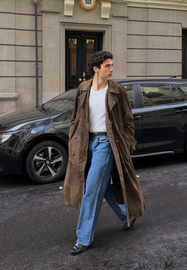 Man wearing brown trench coat, white shirt, and jeans walking on city street past black car and stone building with black door.