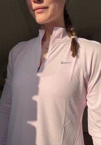 Γυναίκα με ροζ ελαφριά ζακέτα Nike με φερμουάρ και μικρό λογότυπο, με πλεξούδα στα μαλλιά και λεπτά στρωμένα κολιέ.