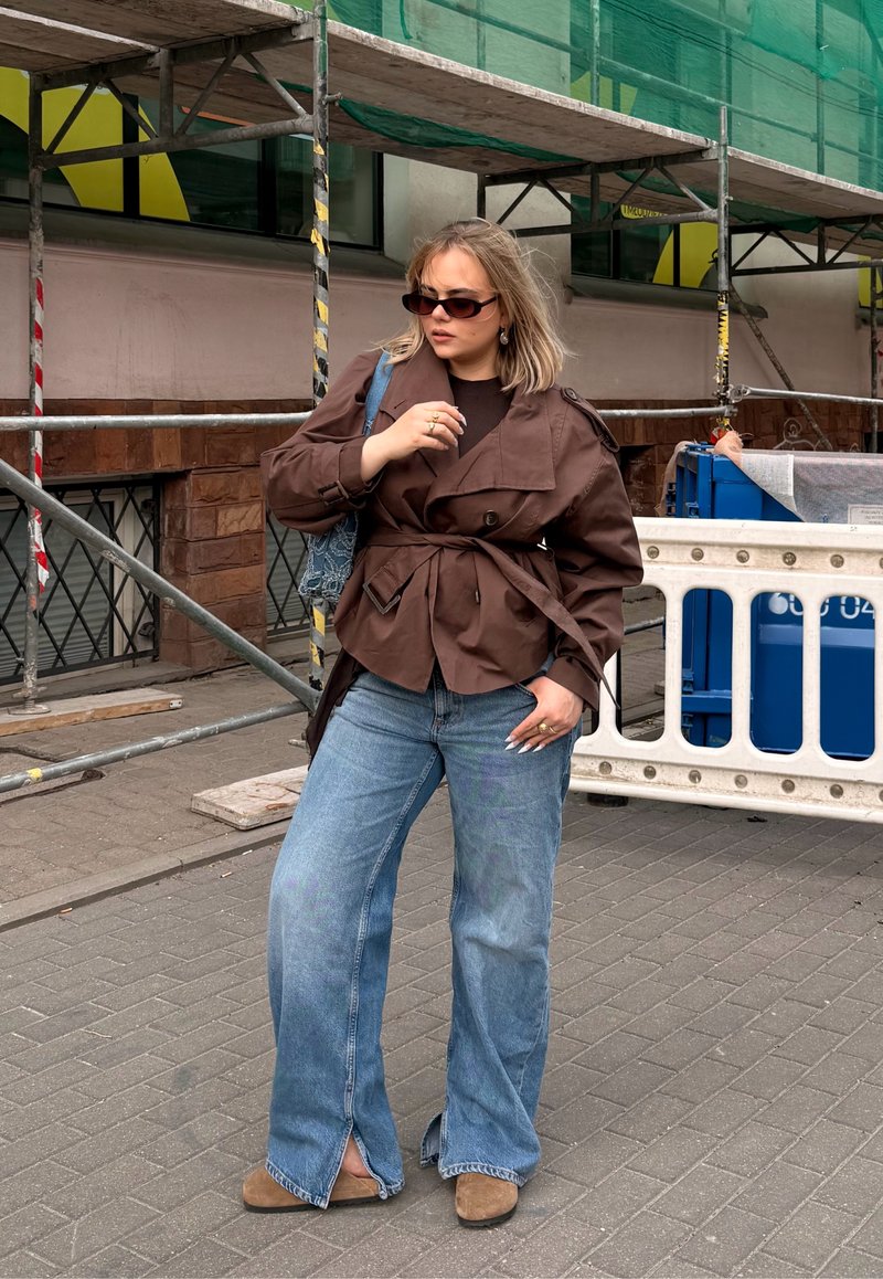 Femme portant une veste marron, un jean large bleu et des lunettes de soleil, debout près d’un échafaudage de construction sur un trottoir de la ville.
