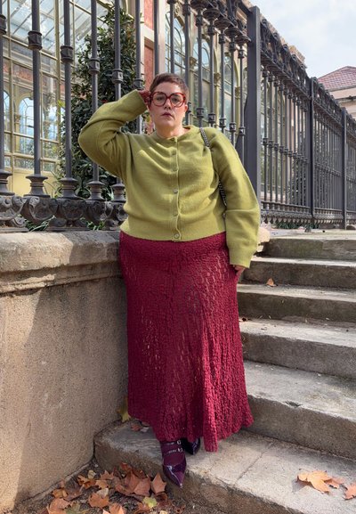 Cárdigan de punto verde, falda maxi de encaje granate y zapatos burdeos de punta fina. Tela texturizada, botones en el cárdigan, de pie sobre escalones de piedra.