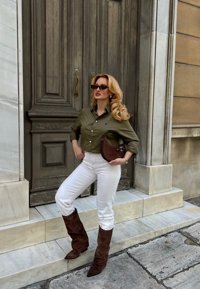 Mujer con gafas de sol posa frente a una gran puerta de madera, vestida con camisa verde, pantalones blancos, botas marrones y sosteniendo un bolso marrón.