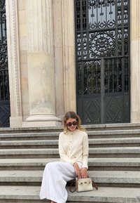 Biele kardigán s textúrovanou látkou, zapínaním na gombíky a dlhými rukávmi; skombinovaný s bielou maxi sukňou a svetlou kabelkou na kamenných schodoch.