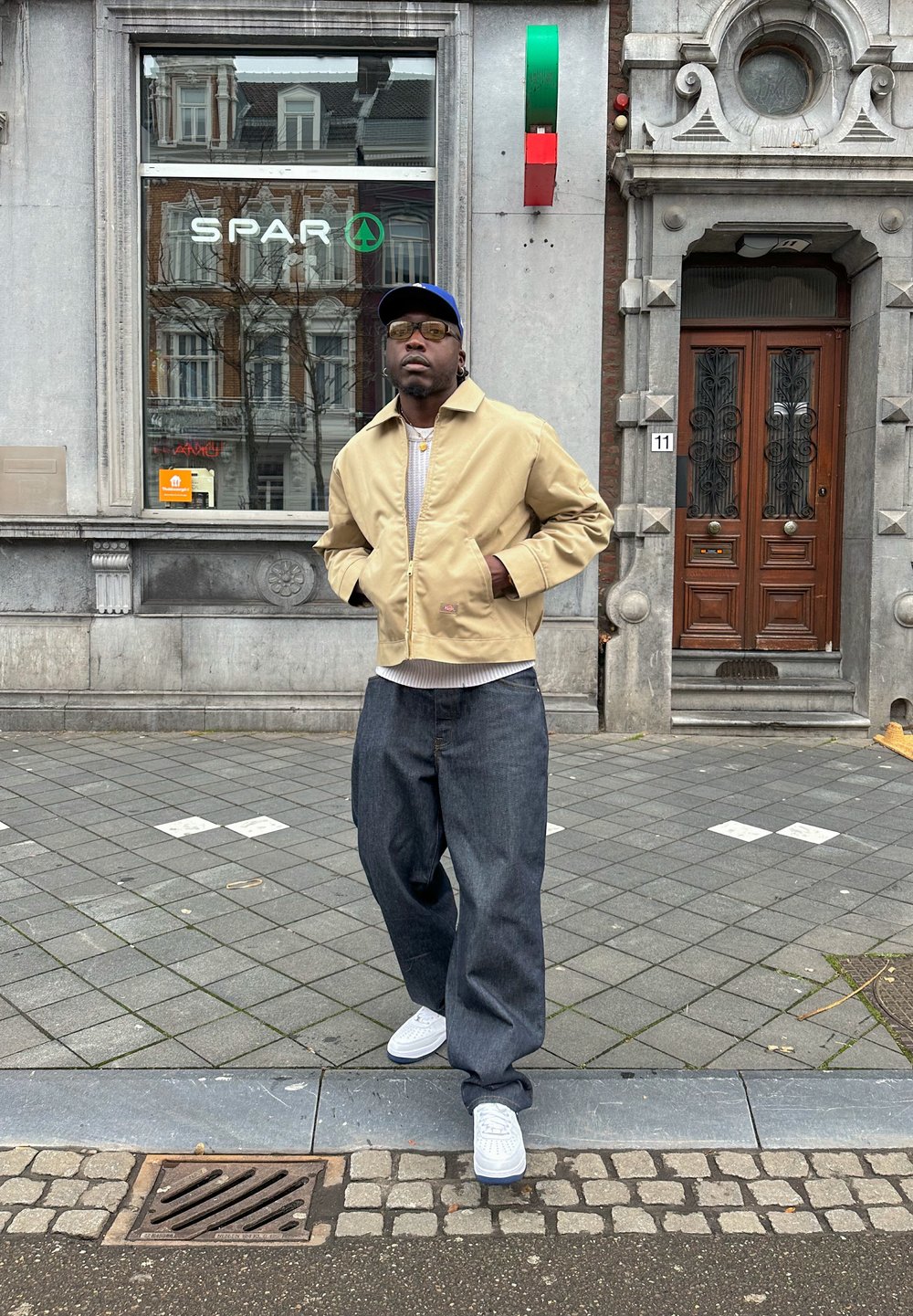 Beige lichtgewicht jas, grijze geribde shirt, losse donkere spijkerbroek en witte sneakers. Accessoires bestaan uit doorzichtige zonnebrillen en gelaagde gouden kettingen.