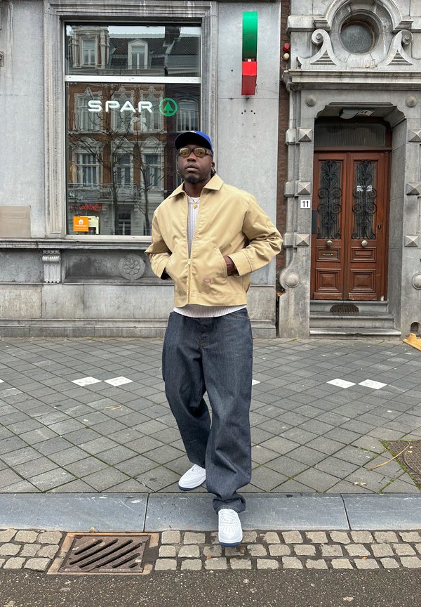 Beige lichtgewicht jas, grijze geribde shirt, losse donkere spijkerbroek en witte sneakers. Accessoires bestaan uit doorzichtige zonnebrillen en gelaagde gouden kettingen.