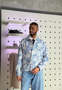 Mann in blauer Batik-Jeansjacke und Jeans steht neben einem weißen Lochblech mit Regalen, die Schuhe und einen hängenden beigen Strickschal halten.