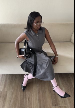 Mujer con gafas sentada en un sofá beige, vistiendo un vestido sin mangas gris, medias rosas, zapatos negros de punta y sosteniendo un bolso negro.